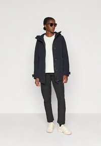 Zwarte waterdichte jas met capuchon, voorzakken en ritsen. Gecombineerd met een wit T-shirt, zwarte broek en beige sneakers.