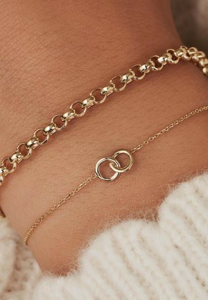 Bracciale in oro con maglie interconnesse, sovrapposto a una delicata catena con due anelli intrecciati. Texture liscia, finitura lucida.