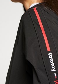 Czarna kurtka z czerwonymi pionowymi paskami i białym logo "tommy" na rękawie; gładka tekstura z sportowym designem.