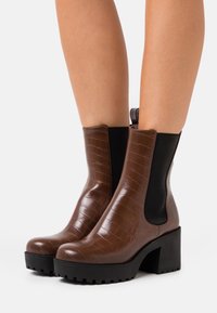 Botines de tobillo en relieve de cocodrilo marrón con paneles elásticos laterales negros y un tacón grueso negro, que presentan una puntera redondeada y una superficie texturizada.