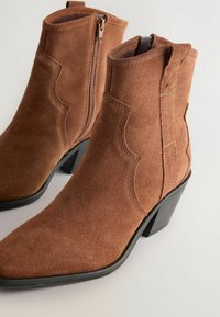 Botas de tobillo de ante marrón con punteras puntiagudas, cremalleras laterales y tacones cuadrados cortos. Presentan detalles de costura en un patrón decorativo.