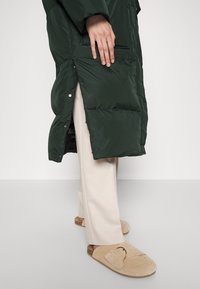 Grønne pufferjakke med et elegant design, der har høj krave, trykknapper og sidelommer. Sammen med beige brede bukser og brune slippers.