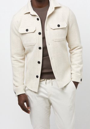 Hombre vistiendo una chaqueta cremosa con textura y botones sobre una camisa marrón, combinada con pantalones blancos con cordón, de pie con una mano en el bolsillo.