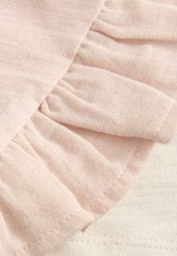 Vaaleanpunainen röyhelöinen kangas pehmeällä, teksturoidulla pinnalla limittää vaaleanbeigeä, sileää kangasta. Röyhelö tuo ulottuvuutta ja yksityiskohtia suunnitteluun.