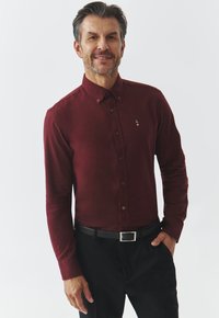 Burgunderfarvet skjorte med lange ærmer og knap-ned krave, der har et lille logo på brystet, parret med sorte bukser og bælte.