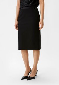 Schwarzer knielanger Bleistiftrock aus glattem Stoff mit einem Bund. Kombiniert mit schwarzen spitzen Absatzschuhen für einen eleganten Look.