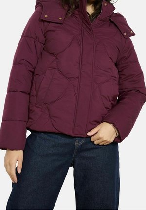 Kobieta w pikowanej, ciemnofioletowej kurtce puchowej z przednimi kieszeniami i odpinanym kapturem, ubrana w ciemnoniebieskie dżinsy.