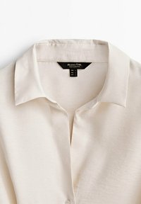 Lichtbeige blouse met kraag van gladde stof met een subtiele glans. Voorzien van een knoopsluiting en een zichtbare label binnenin de kraag.