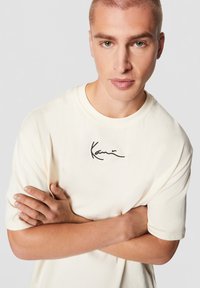 Jeune homme aux cheveux courts et aux yeux verts portant un t-shirt crème avec un logo signature noir, bras croisés devant un fond uni.
