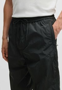 Musta kevyet housut, joissa on joustava vyötärö, nyöri, sivutaskut ja pieni logo-ompelu reidessä. Rakenteellinen kangas.