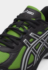 Κοντινό πλάνο του πράσινου και μαύρου αθλητικού παπουτσιού Asics που αναδεικνύει τα κορδόνια, τη λεπτομερή ραφή και το λογότυπο στη γλώσσα και στην πλευρική επιφάνεια.