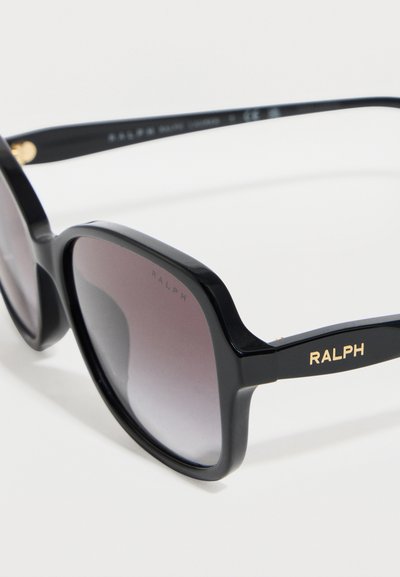 Μαύρα γυαλιά ηλίου με τετράγωνο πλαίσιο, φακούς με σκούρα βαθμιδωτή απόχρωση και χρυσό λογότυπο "RALPH" στο πλαϊνό μέρος, τοποθετημένα πάνω σε λευκή επιφάνεια.
