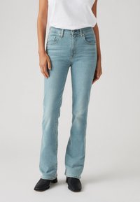 Svetlo modri traper jeans z razširjenim krojem, pet žepov in subtilnim bledkom. Ujemajo se z črnimi gležnjarji na peto.