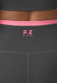Tmavě šedé legíny s růžovým pasem a logem "P.E NATION" v růžovém písmu. Hladká textura se zasazenými švy.