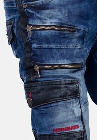 Un pantalon cargo en denim bleu avec de multiples poches zippées, un tissu texturé et des accents foncés contrastants. Marquage brodé en rouge.