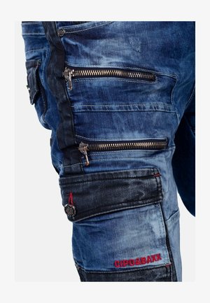 I pantaloni cargo in denim blu presentano numerose tasche con zip, un tessuto testurizzato e accenti scuri a contrasto. Branding ricamato in rosso.