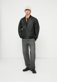 Vīrietis, kurš valkā melnu ādas bumbvedēja jaku, brūnu kreklu ar melnu kaklasaiti, pelēnas džinsas, melnus apavus un krēmkrāsas adītu cepuri, stāvot pret balto fonu.