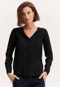 Blusa nera a maniche lunghe con scollatura a V, apertura con bottoni e orlo leggermente curvo. Realizzata in tessuto morbido con una vestibilità comoda e maniche con polsini.