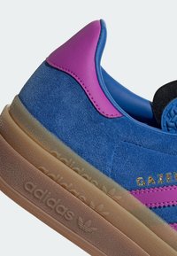 Blauwe suède sneakers met roze accenten, voorzien van een gestructureerde tussenzool en gouden "GAZELLE" letters. Rond gevormd met een gewatteerde kraag.