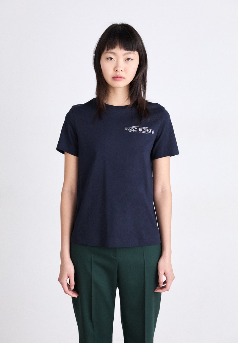 T-shirt en coton bleu marine avec un col rond, des manches courtes et un graphique logo GANT sur la poitrine gauche. Texture lisse et coupe régulière.