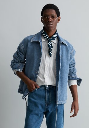 Jeune homme portant de grandes lunettes, une chemise rayée, une écharpe bleu foncé et blanche, une veste en denim bleu et un jean, posant devant un fond gris.
