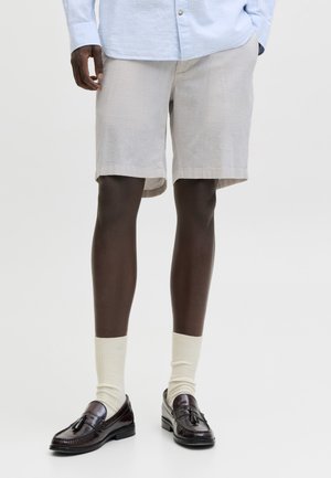 Άτομο που φοράει μπεζ σορτς, ανοιχτό μπλε πουκάμισο με μακριά μαν sleeves, κρεμ κάλτσες και σκούρα καφέ μοκασίνια με φούντες, στέκεται μπροστά σε μονόχρωμο φόντο.