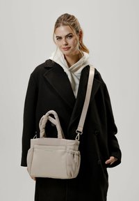 Vaaleanbeige pehmustettu käsilaukku, jossa on kaksinkertaiset kahvat ja säädettävä olkahihna. Pehmeä rakenne ja minimalistinen muotoilu, jossa on sivutaskut ja hillitty logo.