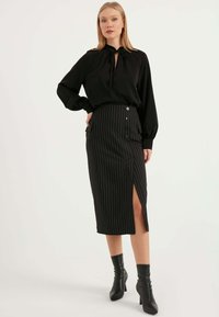 Svart blus med knytning vid halsen, långa ärmar och en svart randig midikjol med knappar och en sidoflik, parat med svarta klackstövlar.