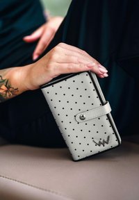 Šedá kožená peněženka s černými puntíky, s klopou na zacvaknutí a stříbrným logem. Hladká textura a obdélníkový tvar.