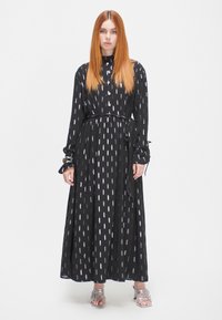 Schwarzes Maxikleid mit durchgeknöpftem Vorderteil, einem Bindegürtel und einem Muster aus vertikalen weißen Ovals. Lange Ärmel mit Knopfdetails, getragen mit silbernen Sandalen.