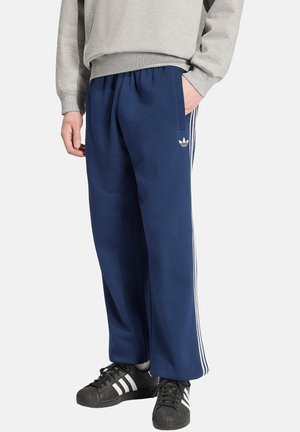 Person iført blå Adidas træningsbukser med hvide striber og sorte Adidas sneakers, stående med den ene hånd i lommen.