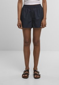 Personne portant un short noir à taille élastique associé à des sandales noires à double bride et un haut blanc sans manches, debout devant un fond gris.