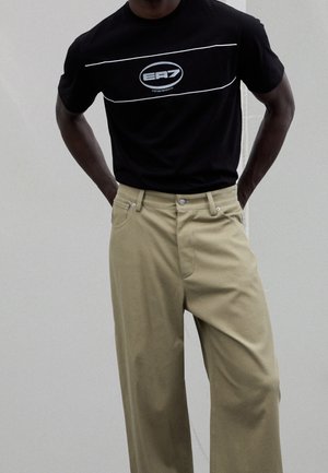 Mand iført en sort EA7 T-shirt med korte ærmer, der er stoppet ned i beige, højttaljede, vidde bukser med hænderne bag ryggen.