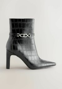 Bottines noires en cuir à motif crocodile, présentant un bout pointu, un talon aiguille et un accent en chaîne argentée autour de la cheville.