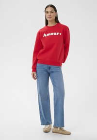 Femme debout portant un sweat-shirt rouge avec le texte "Amour", un jean bleu clair et des baskets beiges, sur un fond blanc uni.