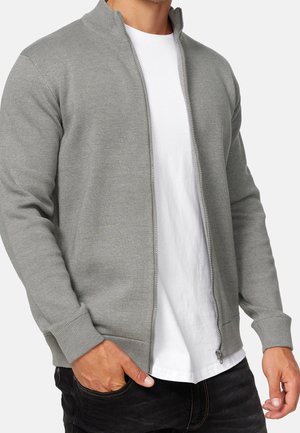 Homme portant un pull zippé gris clair par-dessus un t-shirt blanc avec un pantalon noir, une main dans la poche, sur un fond uni.