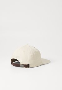 Gorra de algodón beige con visera curvada, correa ajustable en marrón y hebilla de metal en la parte trasera. Presenta ojales cosidos para ventilación.