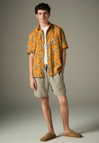 Kurzarm, orangefarbenes Hemd mit Knopfleiste und Palmenmotiv, kombiniert mit hellgrauen Leinen-Shorts und braunen Slippern.