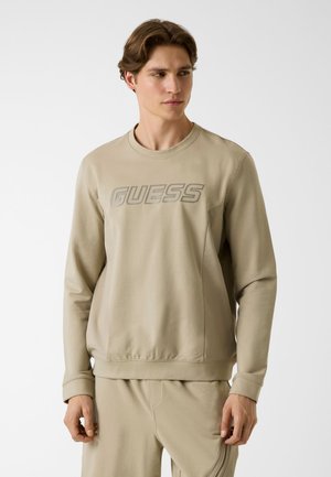 Giovane uomo che indossa una felpa beige e pantaloni coordinati, la felpa presenta il logo "GUESS" sul petto, guarda leggermente di lato.