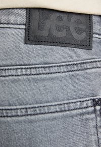 Jean en denim gris avec une étiquette texturée noire portant la marque "Lee". Coutures visibles le long des poches et de la taille.