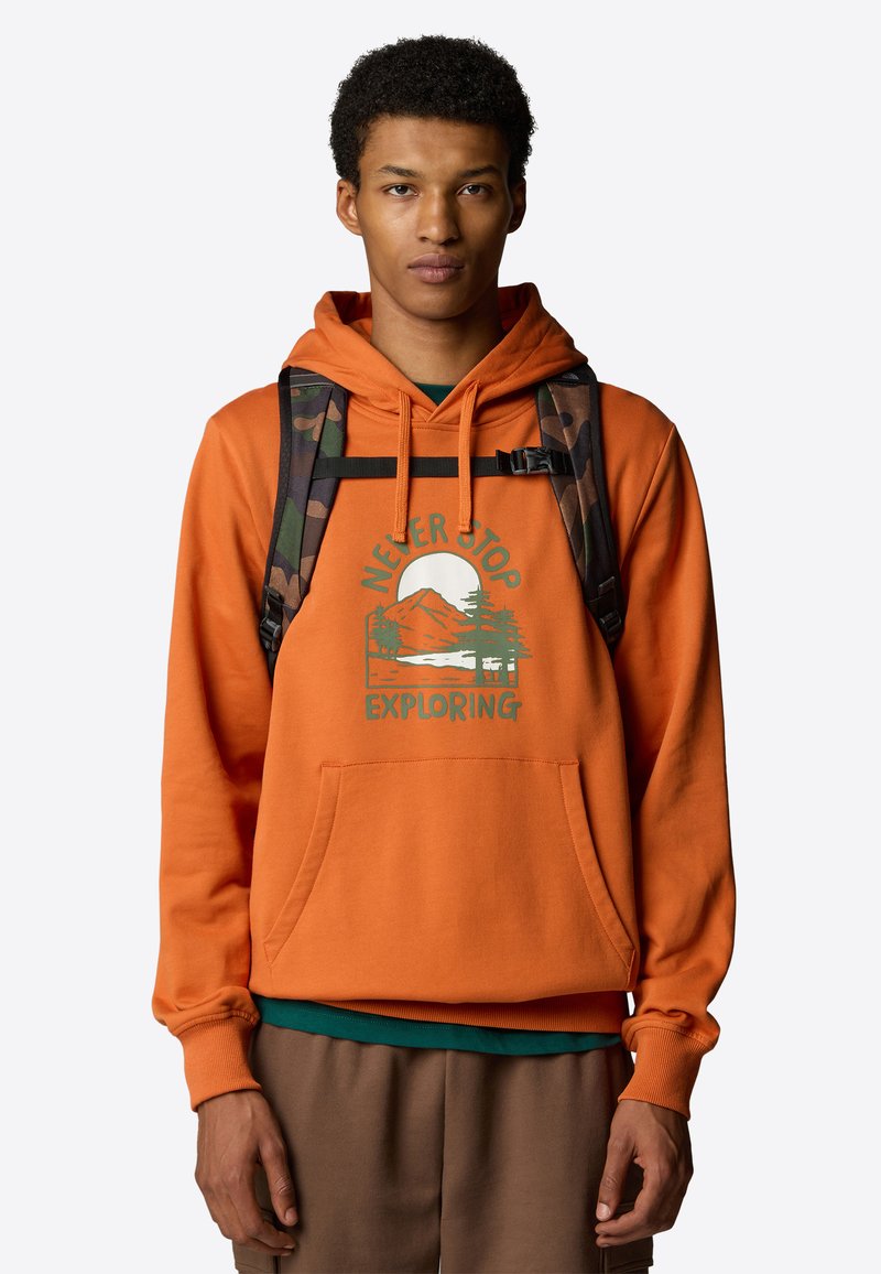 Oranžová mikina s kapucí a přední kapsou, s grafickým potiskem hor a stromů. V kombinaci se zeleným tričkem a hnědými kalhotami.
