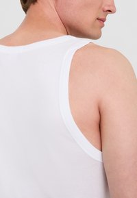 Vit ärmlös tanktop i bomull, rund halsringning, breda ärmhål, slät textur, synlig sömdetalj vid kanterna.