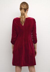 Röd sammet klänning med trekvartsärmar, rundad halsringning och utställd design. Slät textur, synliga sömmar längs baksidan.