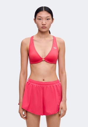 Ung kvinde med sort hår i en ponytail iført en rød sports-bh og matchende atletikshorts, stående mod en ensartet lys baggrund.