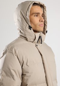 Giacca puffer beige con cappuccio, caratterizzata da una trama di tessuto liscia, chiusure a scatto e cerniera, e cordini regolabili sul cappuccio.
