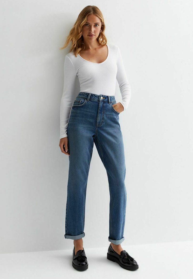 Top bianco leggero a maniche lunghe abbinato a jeans blu in denim a vita alta, con una vestibilità rilassata e polsini arrotolati. Mocassini neri completano il look.