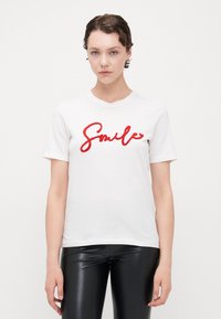 Hvit bomull T-skjorte med korte ermer som har rød brodert "Smile" tekst i midten. Kombinert med svarte blanke bukser.