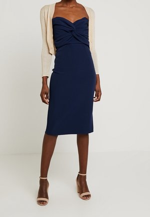 Frau trägt ein marineblaues trägerloses Midikleid mit einer gedrehten Vorderseite, einen beigen kurzen Cardigan und beige Stöckelschuhe mit Knöchelriemen.