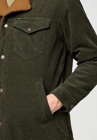 Chaqueta de pana verde con cuello de forro polar color beige, dos bolsillos en el pecho y botones de metal. Presenta canalones verticales y detalles de costura en contraste.