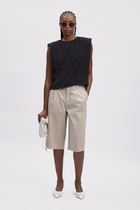 Sort ærmeløs top med plisseret design, beige skræddersyede shorts, hvide spidse flade sko og en sølvfarvet tekstureret taske.